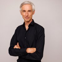 Portret van Hans Hermans. Hij staat voor een neutrale beige achtergrond met zijn armen over elkaar, lacht vriendelijk en draagt een zwarte blouse.