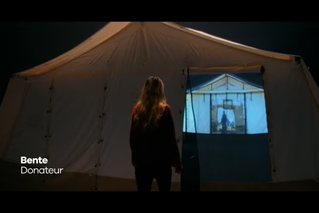 Een vrouw genaamd Bente, aangeduid als donateur, staat voor een grote witte tent waarop een beeld van een medische noodsituatie geprojecteerd wordt.