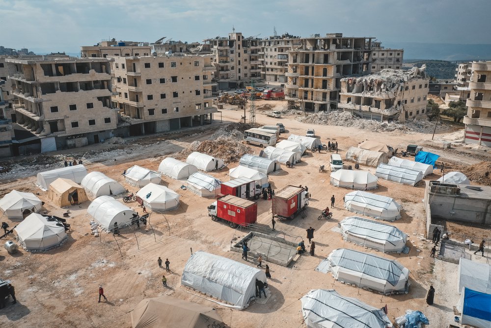 Een luchtfoto van een zandplein in Syrië tussen door de aardbeving verwoeste gebouwen waar witte medische tenten zijn opgezet