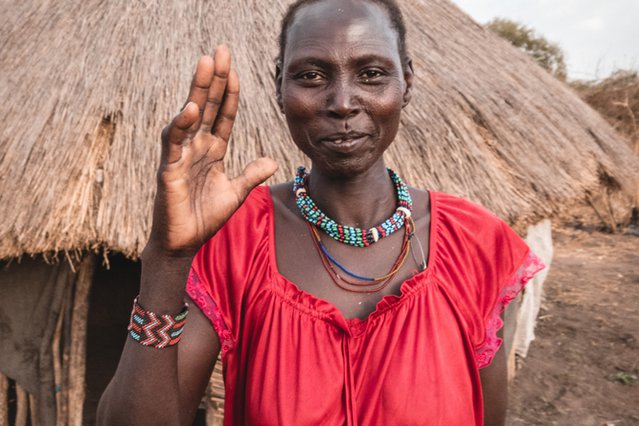 Een vrouw genaamd Roda James zwaait naar de camera. Ze staat voor een huis met rieten dak in haar dorp Labarab in Zuid Soedan.