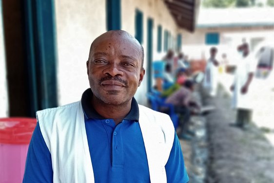 Verpleegkundigbegeleider Denis Mudake staat in een wit Artsen zonder Grenzen jasje voor een gebouw van het Kiseguru gezondheidscentrum