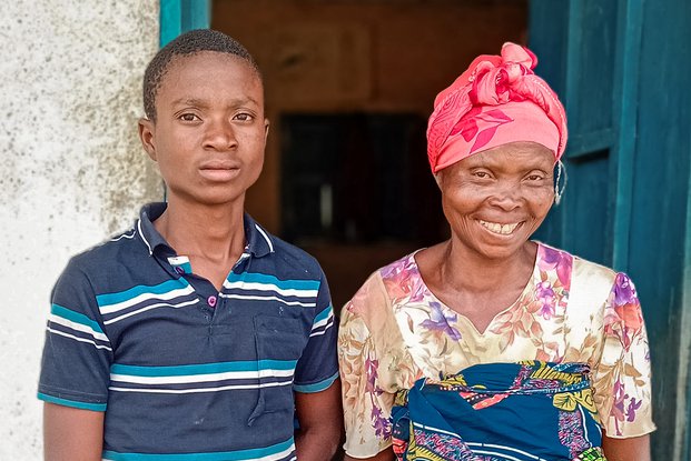 De zestienjarige Dieu-est-bon staat naast zijn moeder voor hun huis. Zijn moeder heeft een grote lach op haar gezicht.